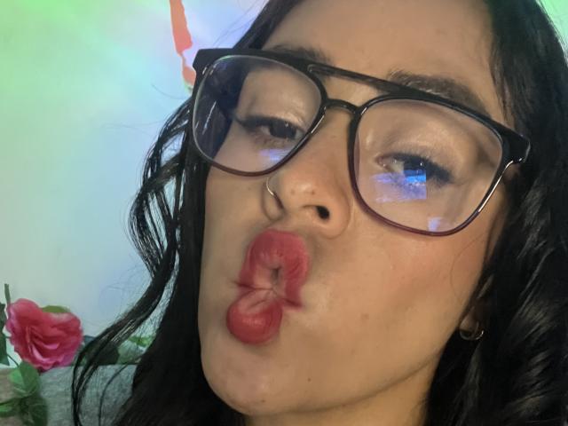 StellaBeckker with pouty red lips, black wavy hair, and black glasses, making a kissy face, nose ring visible, soft green-blue reflection on glasses. Rose in bottom left.