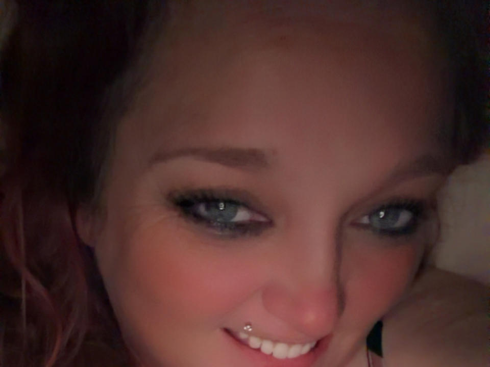 El rostro de Twisttedcandy, con ojos azules, delineador negro y un piercing plateado en la nariz, sonríe ligeramente. Su cabello rosado-rojizo es parcialmente visible. La imagen es un selfie borroso..