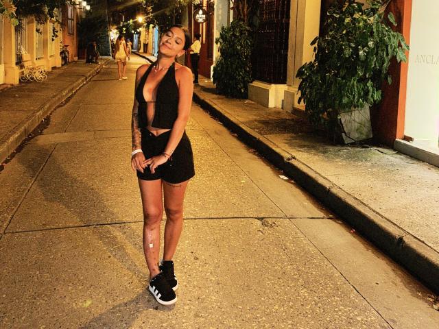 ThaliaAres stands on a nighttime street, wearing a black sports bra, shorts, and sneakers, with tattoos on her arm and a confident smile, her tits slightly visible.