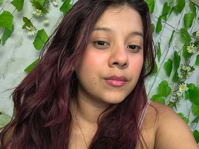 CoralineHale with dark red hair, nose ring, pink lipstick, slight pout, small moles on cheek, wearing spaghetti strap top, green leafy background with white flowers.