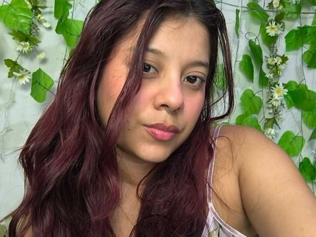 CoralineHale has dark red, wavy hair, a nose ring, and pink lipstick. She's looking at the camera with a slight, confident smile, showing her cute face.