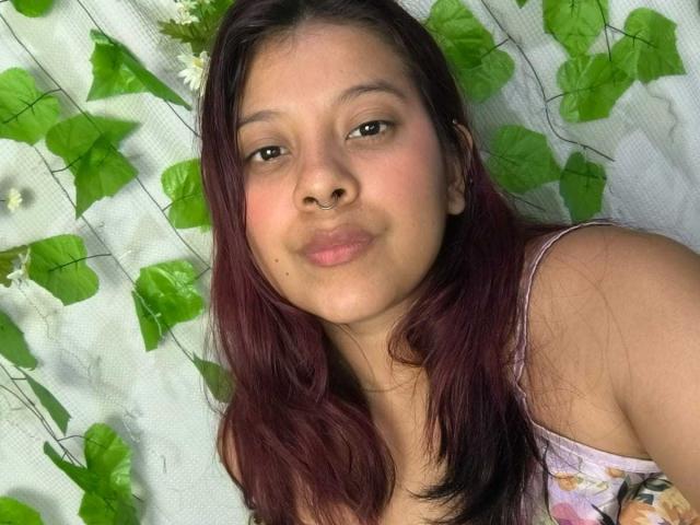 CoralineHale has light brown skin, dark brown eyes, and red-purple hair. She's pouting with her pink floral dress visible, showing small tits. Green leafy background.