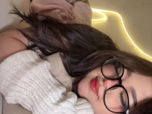 Meagar3 with long brown hair, glasses, and red lipstick, sleeping with a white sweater, her soft tits barely visible under the blanket. Neon light in the background.