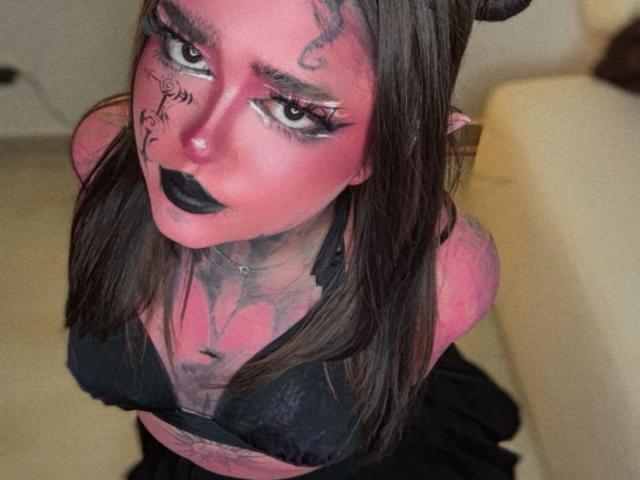 Meagar3 with pink face makeup, black lipstick, and tattoos, wearing a black lace bra, has brown hair, and devil horns, looking up with large eyes and dark eyeliner.