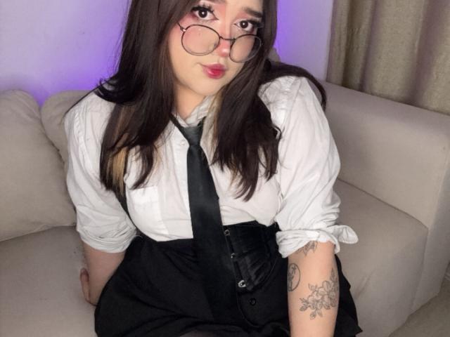Megar3 with glasses, white shirt, black tie, and black pants, sitting on a couch, showing a tattoo on her left arm, with medium tits and dark brown hair.