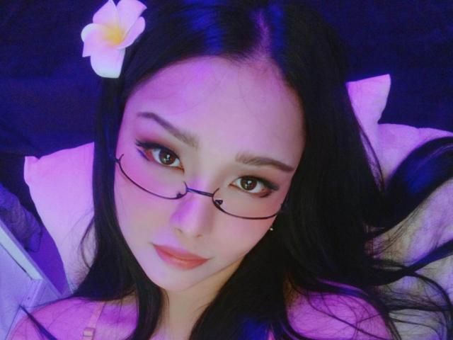 JenieMistress with black hair, glasses, and a white flower in her hair, lying down with purple and blue lighting, looking up with a cute expression, showing her tits.