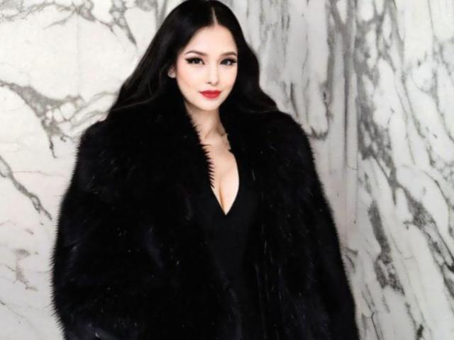 JenieMistress with long black hair, red lipstick, and black fur coat, showing her tits in a deep V-neck black dress, standing in front of marble walls.