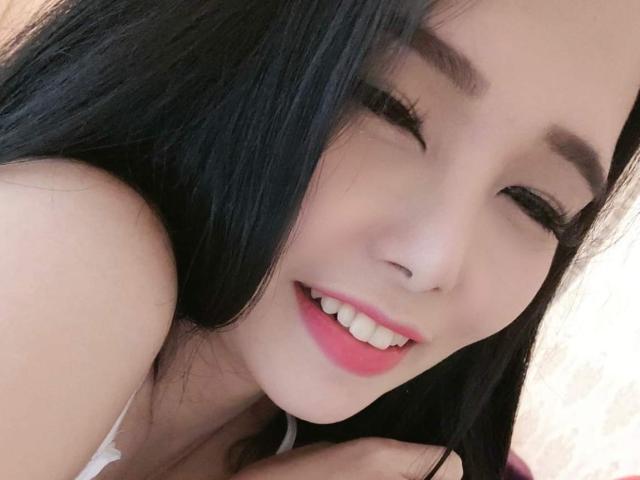 JenieMistress has long black hair, a cute smile with pink lips, and is winking. Her small nose and white teeth are visible. She's wearing a white strap.