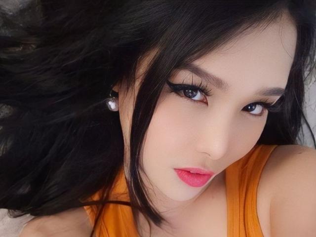 JenieMistress has long black hair, blue eyes with heavy eyeliner, pink lipstick, and wears an orange top. She's lying down, looking up with a slight pout.