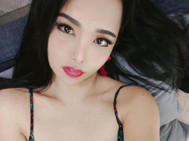 JenieMistress with long black hair, red lipstick, and dark eyeliner, lying down, wearing a colorful, thin-strapped top, showing her small, perky tits and soft, fair skin.