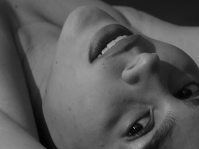 Le visage de RossaHot sur une photo en noir et blanc, allongée, les yeux levés, les lèvres légèrement entrouvertes, exprimant une expression détendue. Son nez est proéminent et ses lèvres sont pulpeuses..