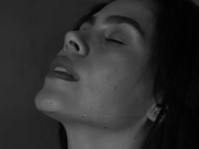 RossaHot's face in a black-and-white photo, eyes closed, head tilted back, mouth slightly open, showing a bit of her teeth, with a small earring in her right ear.