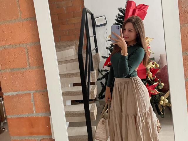 Gabrielafoxxy with long brown hair, green long-sleeve top, and beige pleated skirt, holding phone, mirror selfie, red bow, Christmas tree, beige stairs, and brick wall.