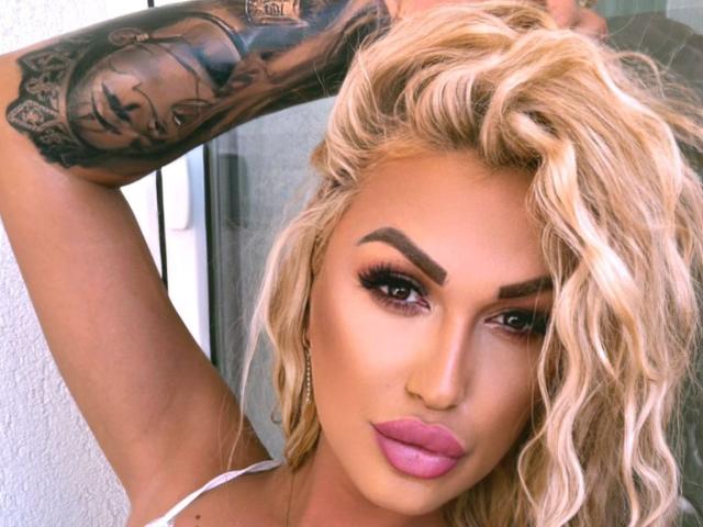 Blonde NicolSNaya with wavy hair, tattooed arm, bold eyebrows, long eyelashes, pink lipstick, and a sexy gaze, raising her arm, showing off her tits slightly.