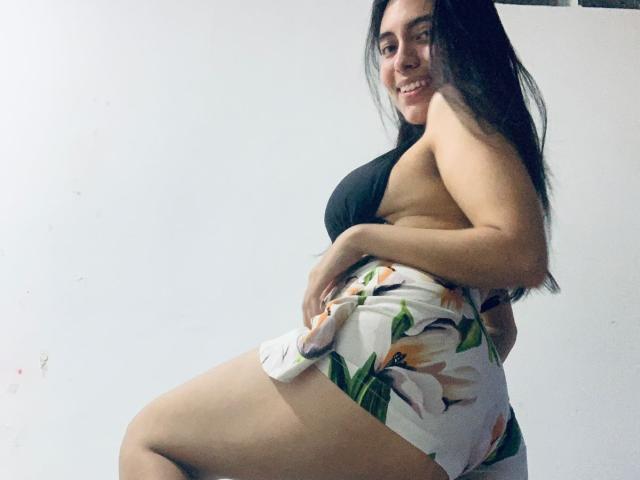 EmilySexyyx with long black hair, smiling, wearing a black bra and white floral shorts, showing her ass and curvy body, standing sideways, looking over her shoulder.