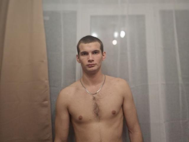 Shirtless Noctyve with a short buzzcut, wearing a silver chain necklace, standing in a bathroom with a beige curtain on the left and translucent curtains in the background.