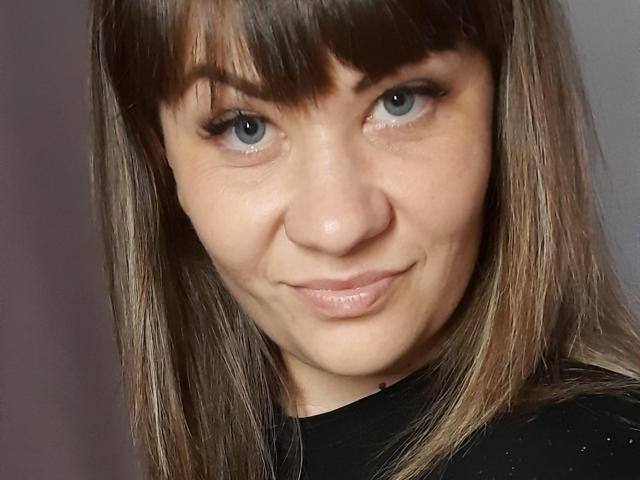 EmilyLunar with brown bangs, blue eyes, and a slight smile, showing her nose and shiny pink lips, wearing a black top, in a selfie.