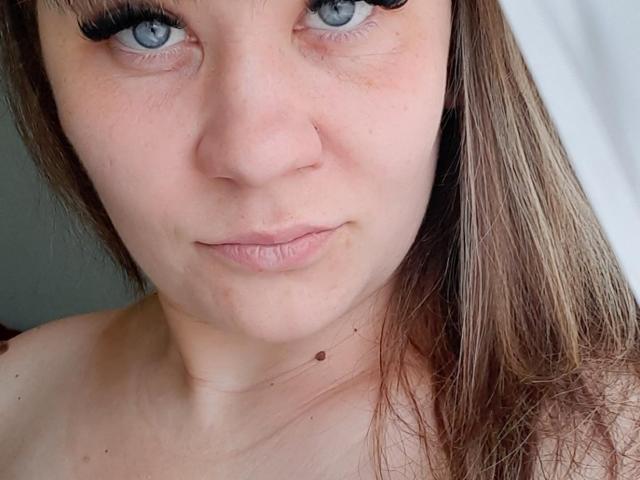 EmilyLunar has light skin, blue eyes, and brown hair with bangs. She's looking at the camera with a slight smirk, wearing long black eyelashes. Her tits are not visible.
