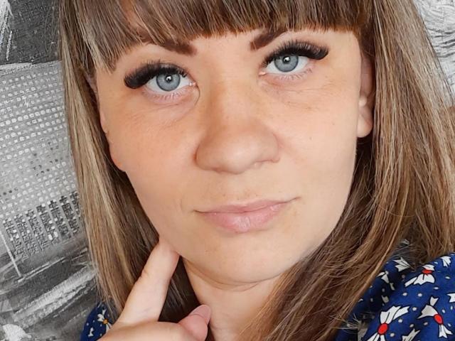 EmilyLunar has blue eyes, brown bangs, and wears long black eyelashes. She's touching her chin with her finger, wearing a blue floral shirt, and has a slight smile.