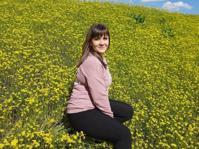 EmilyLunar with medium tits, smiling, kneeling in a yellow flower field, wearing a pink shirt and black pants, brown hair, blue sky, and white clouds in the background.