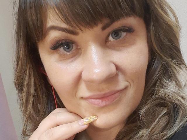 EmilyLunar with brown wavy hair, blue eyes, and gold glitter nail, touching her chin, smiling slightly, wearing red earbuds, and looking at the camera.