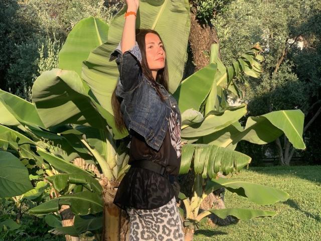 LunaGalaxy with long dark hair, wearing a denim jacket and leopard print skirt, stretching arms up, standing in front of large green banana leaves in a sunlit garden.