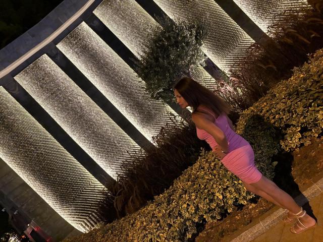 LunaGalaxy with long brown hair, pink dress, and pink heels, standing with ass slightly raised, looking at illuminated wall, in a well-lit garden at night.