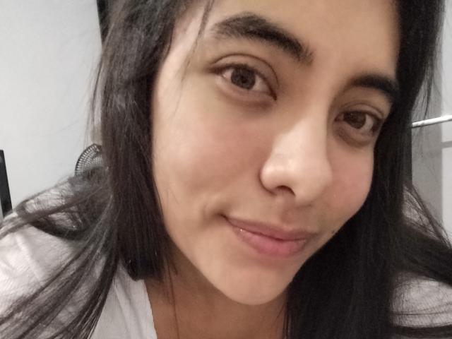 EmilySexyyx has light brown skin, dark brown eyes, and black straight hair. She's smiling with pink lips, wearing a white shirt, and has a slight pimple on her cheek.