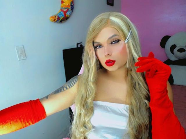 Kimberllys has long, wavy blonde hair, wearing a white strapless top and bright red gloves. She has bold red lipstick, heavy eyeliner, and a tattoo on her right arm. Her right hand is raised, touching her hair. The background includes a colorful wall decoration and a large panda plushie. The image has a bright, colorful style with clear contrasts.