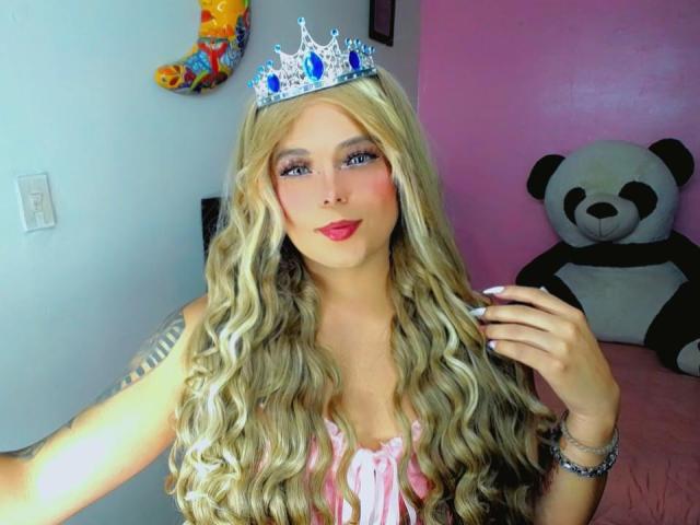 Kimberllys has long, wavy blonde hair and wears a silver tiara with blue gems. She has blue eyes, pink blush, and red lipstick. Her tattooed arm is partially visible. She wears a pink, ruffled top. Her right hand is near her face. In the background, a large black-and-white panda plushie is on a pink bed. A colorful balloon is on the left wall. The room has pink and white walls. The image is bright and focused on Kimberllys.