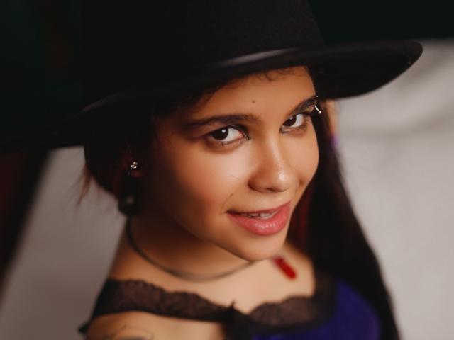 AshGraven with dark eyes, brown skin, and a small nose ring, wearing a black hat and a lacy top, smiling at the camera with a subtle smirk.