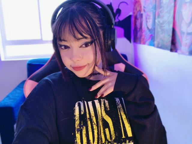 Arieyaara with black headphones, bangs, and dark eyes, wearing a black "PINK" sweatshirt, sitting in a chair, smiling with pink lipstick, right hand on chest, blue room.