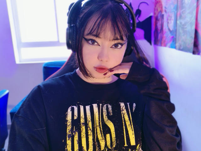 Arieyaara mit dunkelbraunen Haaren, die einen schwarzen "PUNKY"-Pullover, schwarze Kopfhörer und lila Augen-Make-up trägt, lehnt ihr Kinn in ihre Hand und hat ein Nasenpiercing..