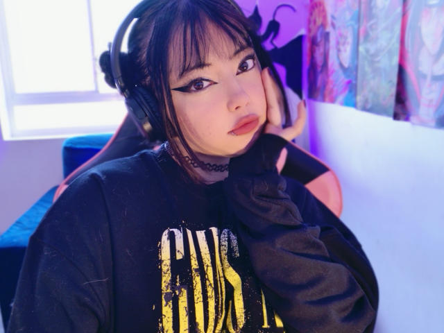 Arieyaara, a young woman with pale skin and dark bangs, wears a black choker and large headphones. She has thick, winged eyeliner and pink lipstick. She's in a black sweatshirt with a yellow and black abstract design. Her right hand touches her cheek. The room has purple and blue lighting, with a colorful wall to the right. The image has a soft, slightly blurry texture. Her expression is neutral, with a slight, relaxed pout.