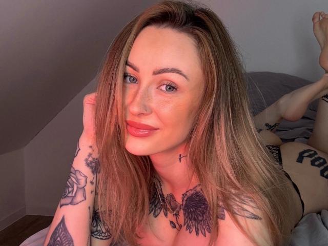 Meltattt-hot with long brown hair, blue eyes, and red lipstick, lying on a bed, showing tattoos on arms and chest, smiling with a hand in her hair, legs up.