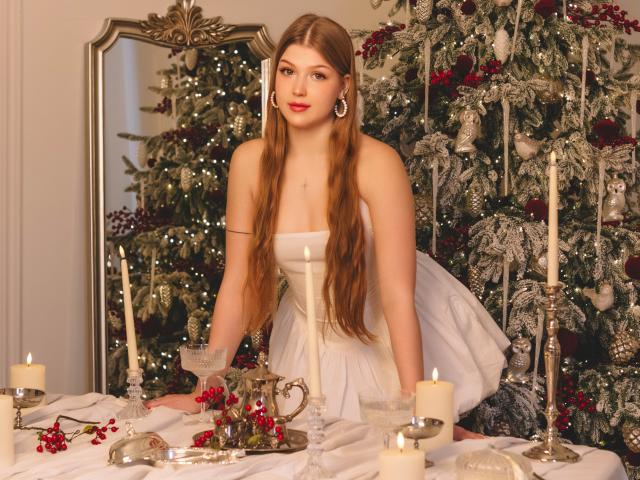 StellaLane, con cabello castaño largo, vestido blanco y lápiz labial rojo, arrodillada sobre una mesa con velas y bayas rojas, mostrando sus senos, en un ambiente navideño..