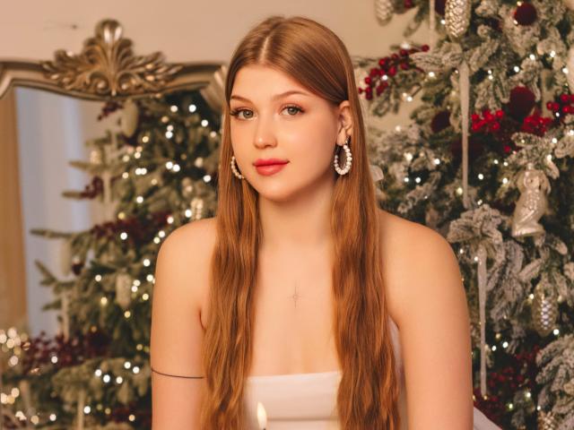StellaLane has long, straight, red-brown hair, red lipstick, and is wearing white earrings, looking forward with a slight smile. Her tits are small and she's wearing a strapless white top.