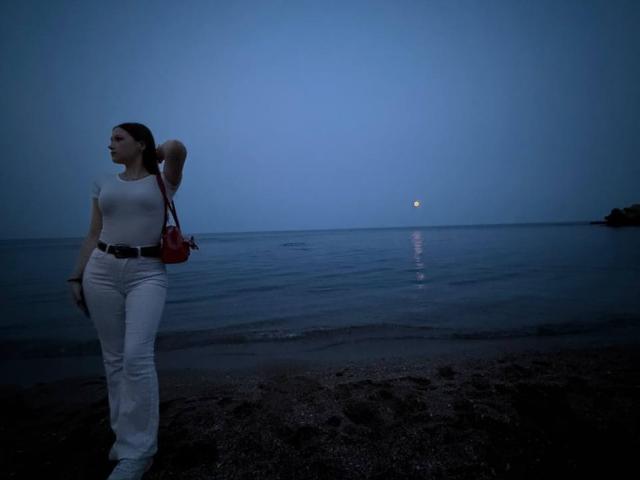 Bubochka with tits in a white shirt, white pants, and red bag, standing on a dark beach at dusk, looking at the moon over the water.