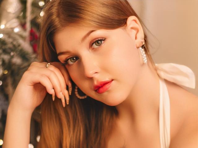 StellaLane has light brown hair, green eyes, red lipstick, and is wearing a white halter top with pearl earrings. She's looking at the camera with a slight smile.