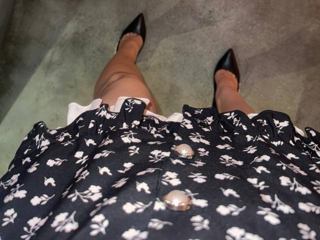 Bubochka's black floral dress with white buttons, showing her legs and black pointed-toe shoes, taken from a downward angle. Her dress is ruffled at the skirt.
