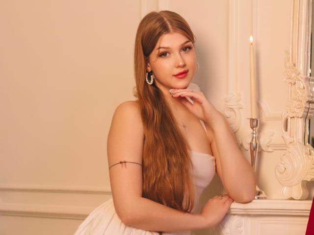 StellaLane, com cabelos castanhos longos, batom vermelho, vestido branco, brincos de pérola, tatuagem no braço, queixo apoiado na mão, vela à direita, sorriso suave, pele clara, olhando para a câmera..