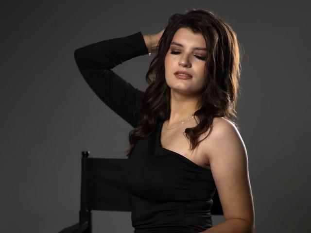 LauraLee69 with wavy brown hair, wearing a black, one-shoulder top, has her hand in her hair, looking slightly down, with a soft, confident expression.