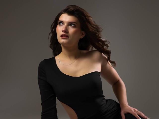 LauraLee69 with wavy brown hair, wearing a black, one-shoulder dress, showing cutout sides, looking up with a slightly open mouth, and a thin necklace.