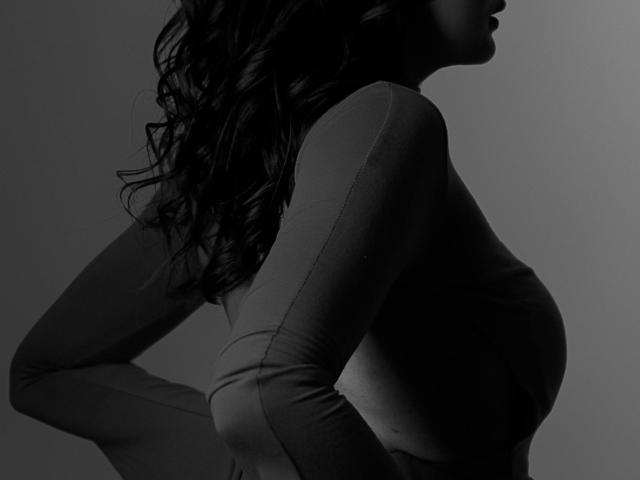 Silhouetted black-and-white photo of LauraLee69 with wavy dark hair, wearing a tight long-sleeve top, showing her small tits and subtle curves, arms raised behind her.