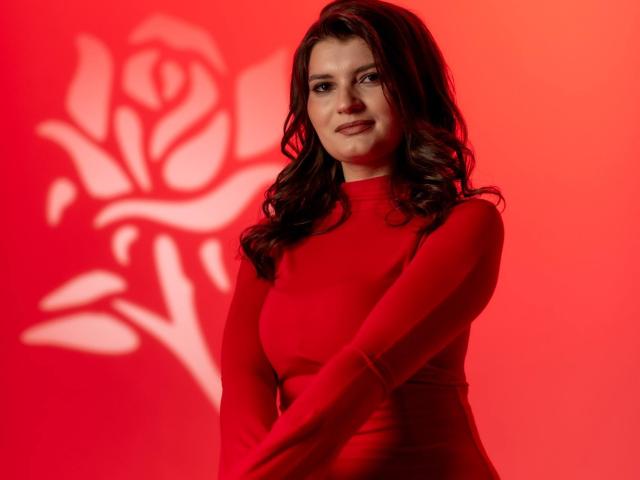 LauraLee69 with wavy brown hair, wearing a red long-sleeve top, standing with arms crossed, smiling slightly, red rose shadow on the wall, red background.