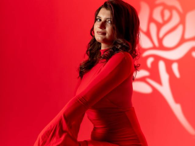 LauraLee69 with wavy brown hair, wearing a tight red dress, sitting against a red wall with a white rose shadow, looking slightly to the side with a subtle smile.