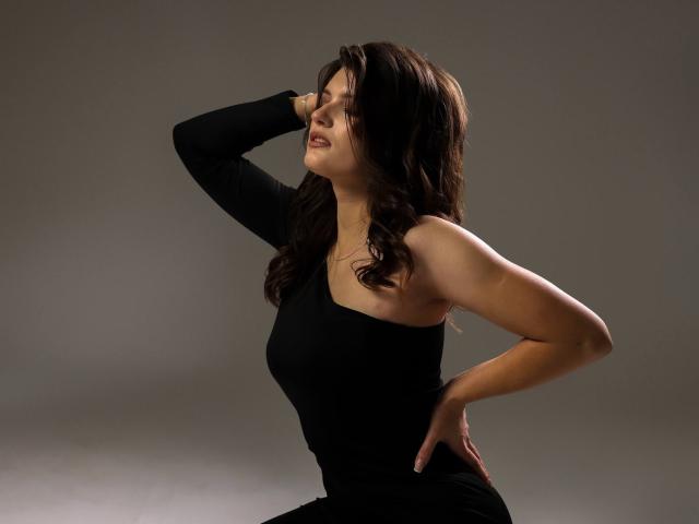 LauraLee69 with wavy brown hair, eyes closed, wearing a black one-shoulder dress, hand on hip, other arm raised, in a provocative, confident pose.