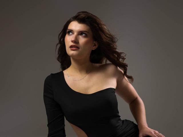 LauraLee69 with wavy brown hair, wearing a black, one-shoulder dress, looking up with a slight pout, her tits subtly outlined, gray background.