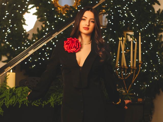 ChristinaBliss with long dark hair, black deep V blouse, red rose on chest, standing, holding plants, candleholder with five candles, warm lighting, subtle smile, gold necklace.