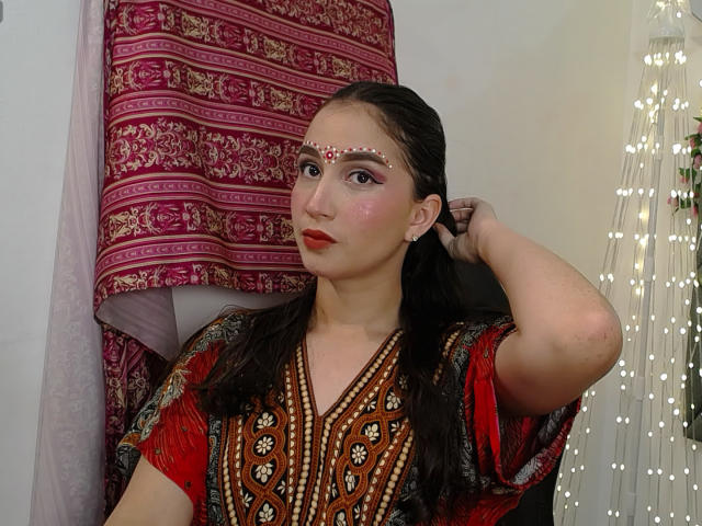 JalimaMona with brown eyes, pink blush, and red lipstick, wearing a beaded red dress, adorned with white facial gems, dark wet hair, hand in hair, string lights.